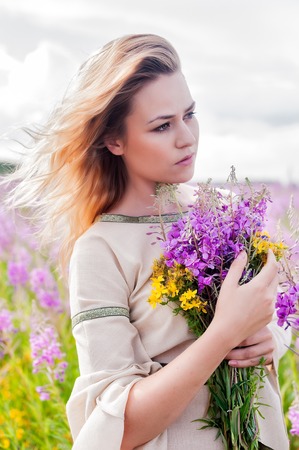 Beautiful woman in traditional dress relaxes with bunch of flowers on fireweed meadowの写真素材