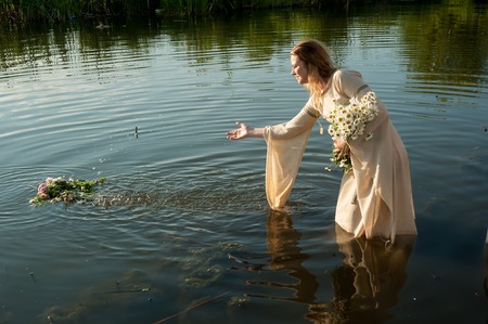 Pretty young woman in traditional dress puts wreath in water of lake. Russian traditional Ivan Kupala holiday celebrationの写真素材