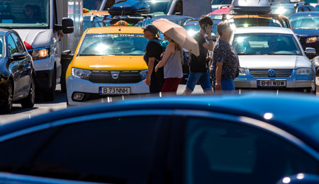 Bucharest, Romania - August 07, 2019: Two women tourists wearing cap and parasol are crossing the street between cars in heavy traffic on a hot day in downtown Bucharest.のeditorial素材