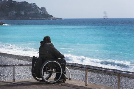 Nice, France, March 2019. A girl in a wheelchair sits on the shores of the azure sea. Beautiful blue...
