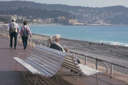 Nice, France, March 2019. A mature, gray-haired beautiful couple: a man and a woman are sitting on a white bench on the Promenade des Anglais and looking at the azure sea.のeditorial素材