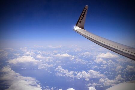 Porto, Portugal, June 2019. White fluffy clouds in the blue sky under the wing of the aircraft of the airline Ryanair. Photos of clouds from the passenger liner porthole. Photo background.のeditorial素材
