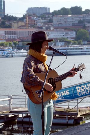 Porto, Portugal, June 2019. A girl in a hat with a guitar sings lyric songs on the embankment of the Douro River in the city of Porto. Street musicians. Romantic trip.のeditorial素材