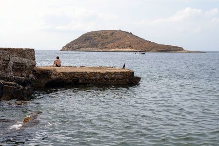 A young guy and a black heron on a stone of Magaluf beach meet the dawn. Loneliness. Sadness. Magaluf, Mallorca, Balearic Islands.のeditorial素材