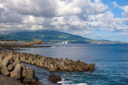 Reinforced concrete tetrapods by the sea in the city of Ponta Delgada protect the coast from waves. Beautiful views of the sea, mountains, clouds and breakwaters. San Miguel Island, Azores, Portugal.の写真素材