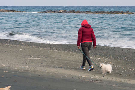 Limassol, Cyprus, February 2020. A girl in a red jacket walks with a little white dog along the seashore. Loneliness. Walk your pet.のeditorial素材