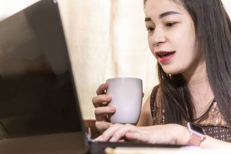 Happy young woman working and learning online from laptop while sitting at the living room, drinking coffee. work from home concept.の写真素材