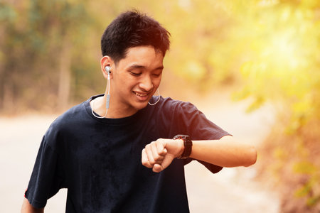 A smiling young man is jogging in the park. He is looking at his watch. He listens to music on his...