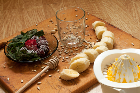 hand squeezes orange juice, banana,frozen strawberries blackberries and seeds vivid smoothie ingredients and blender, juicer, tulips on the background on woodenの写真素材