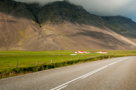 The paved road number 1, a meadow, a farm with red roofs and a mountain masked by clouds in southern Icelandの写真素材