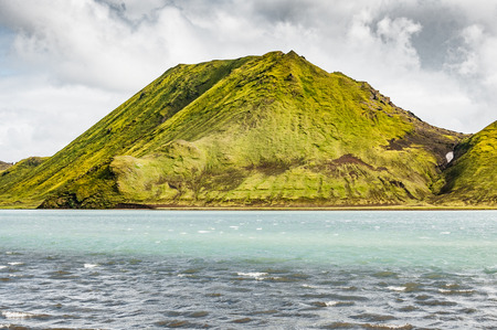 At the edge of the F208 Fjallabak road, the wind creates waves on the surface of a lake. Behind it stands a green mountain covered with grass and lichen.の写真素材