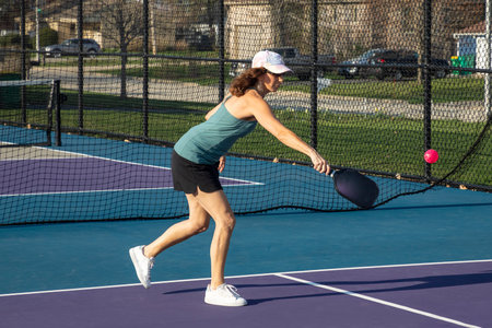 A female pickleball player returns a bright pink ball at the baseline on a dedicated court at a public park.の写真素材