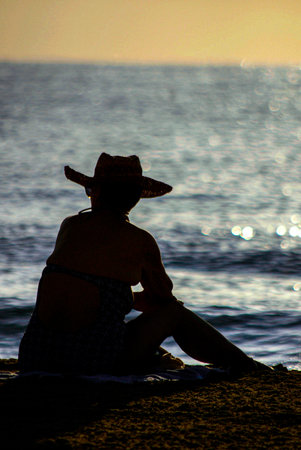 Backlight of a senior woman in a hat sitting on the beach at sunriseの写真素材