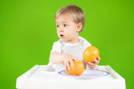 Blond baby sitting in the high chair with oranges in his handsの写真素材