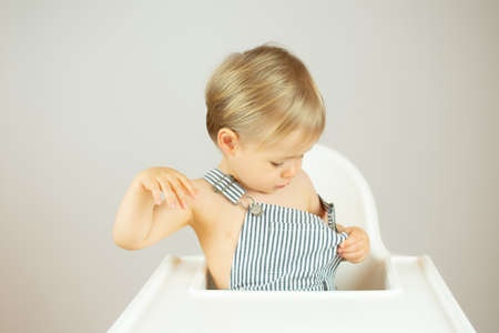 Blond baby with striped overalls sitting in the high chair and smilingの写真素材