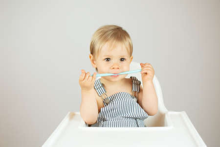 Blond baby with striped overalls sitting in the high chair and with a spoon in his mouth
