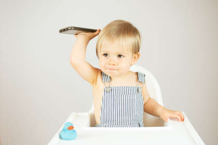 Blond baby with striped overalls sitting in the high chair and picking up the phone