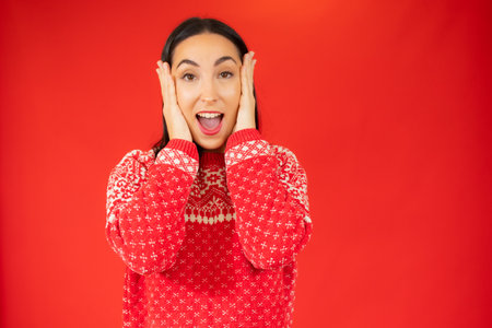 Young woman in winter sweater surprised over red background.