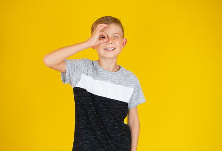 Portrait of handsome boy making okay Gesture with hand on eyes looking through fingers. Beautiful caucasian boy, on yellow background. Happy smiling child showing glasses okay.の写真素材