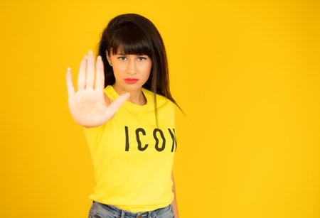 Portrait of a serious young woman standing with outstretched hand showing stop gesture isolated over yellow backgroundの写真素材