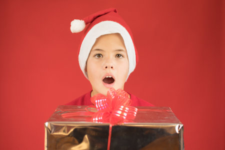 Child in Santa red hat holding Christmas gift in hand with surprise gesture. Christmas concept.