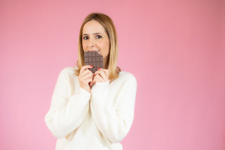 Portrait of a happy young woman biting chocolate bar isolated over pink backgroundの写真素材