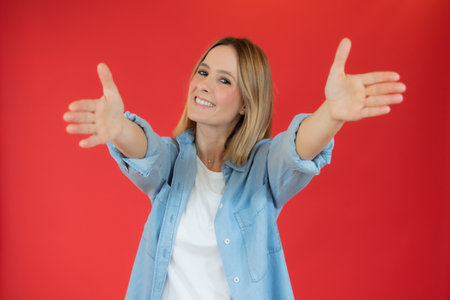 Young beautiful woman wearing denim shirt over red background looking at the camera smiling with open arms for hug. Cheerful expression embracing happiness.の写真素材