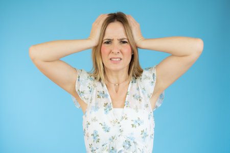 Portrait of young woman with blond hair touching her head and expressing worry isolated over blue...