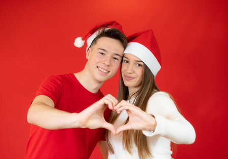 Photo of two charming couple in santa hat making hands fingers heart figure celebrating lovers day isolated red backgroundの写真素材