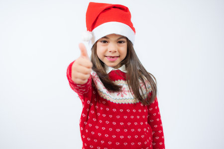 Beautiful smiling little girl in santa hat showing thumb up over white background.の写真素材