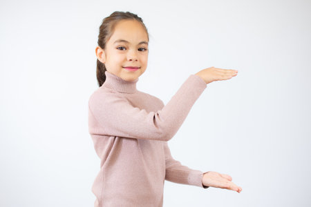 Pretty little girl in pink sweater showing product with her hands over white background.の写真素材
