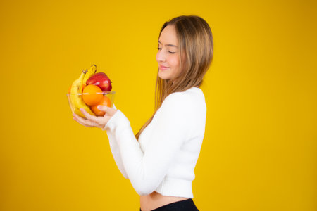 Happy beautiful girl holding bowl of fresh fruits, studio shot, isolated over yellow backgroundの写真素材