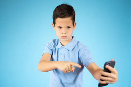 Cute ginger kid with mobile phone on blue studio background, using exciting mobile applications or...