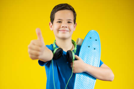 Portrait happy smiling teenager boy wearing a blue shirt and headphones with skateboard in city over yellow backgroundの写真素材