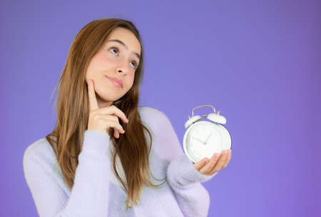 Young woman holding alarm clock over purple isolated background serious face thinking about...