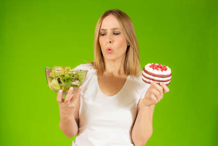 Portrait of a confused young woman choosing between cake and salads, Isolated over green background.