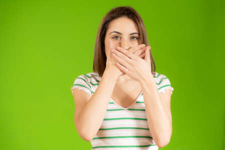 Portrait of beautiful and young woman standing over isolated green background asking to be quiet with hand on lips. Silence and secret concept.の写真素材