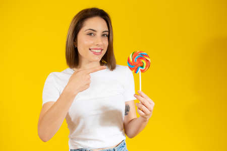 Beautiful young woman holding colorful lollipop candy in hand having fun over yellow background. Sweet food and happiness concept.の写真素材