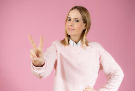 Portrait of a smiling happy woman showing victory sign and looking at camera isolated on the pink backgroundの写真素材