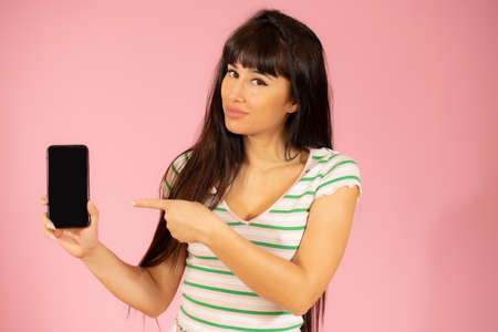 Happy beautiful young woman with long hair showing mobile phone over pink backgroundの写真素材