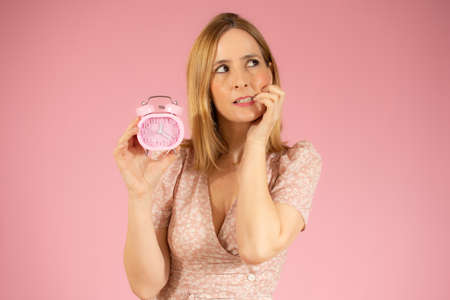 Young excited woman holding classic alarm clock over isolated pink backgroundの写真素材