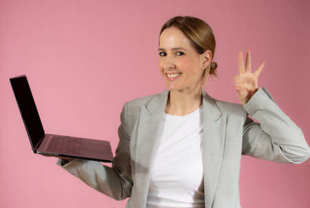 Business young woman holding laptop computer and showing okay sing while looking at the camera over pink backgroundの写真素材