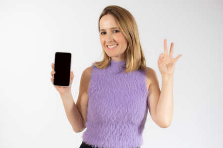 Portrait of a smiling young woman showing blank screen mobile phone with okay sign isolated over white backgroundの写真素材