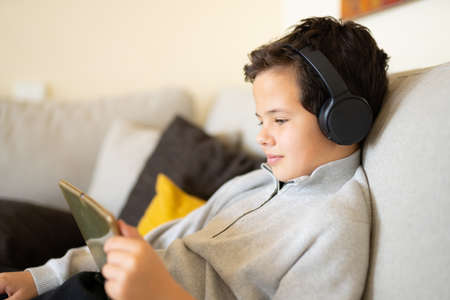 Little smiling boy sitting on sofa and playing on a digital tablet at homeの写真素材