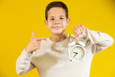 Cute smiling boy pointing with finger an alarm clock isolated over yellow background.