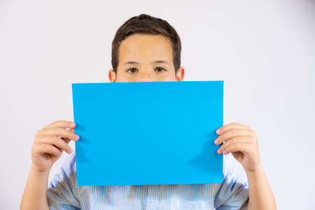 Closeup portrait of a boy holding blue blank paper covering mouth isolated over white backgroundの写真素材
