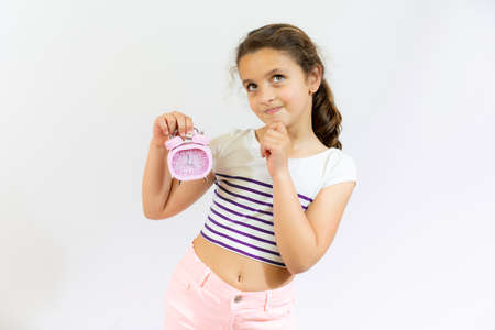 Thoughtful little girl brown-haired child touch chin with finger thinking or considering holding an alarm clock isolated over white background.の写真素材