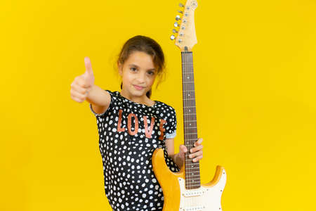 Smiling little girl holding guitar with thumb up over yellow background