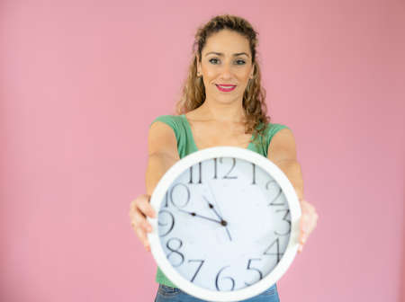 Portrait of a smiling woman in casual t-shirt holding big clock isolated over pink backgroundの写真素材