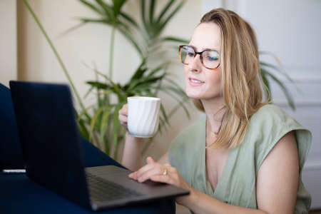 Attractive young woman working on laptop and drinking coffee while sitting on the floor at homeの写真素材